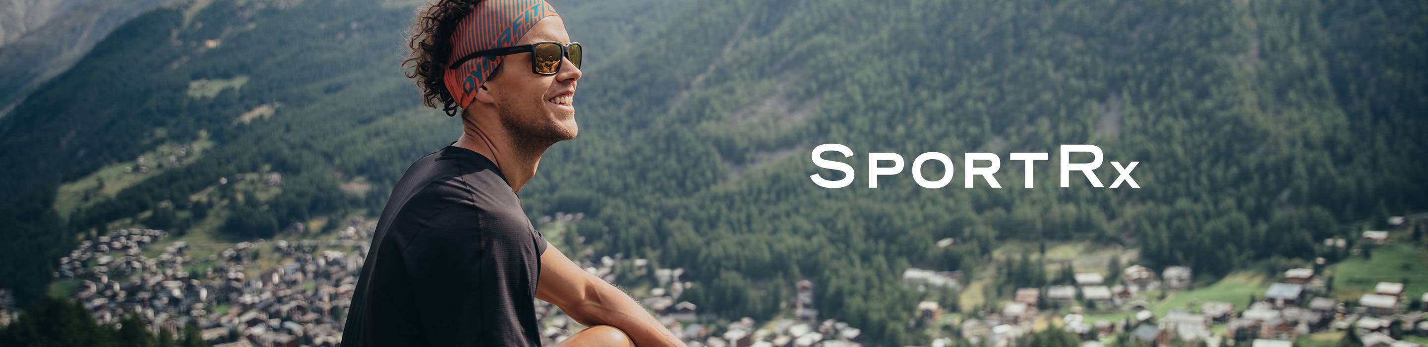A curly haired male hiker wearing SportRx brand sunglasses smiles atop a mountain.